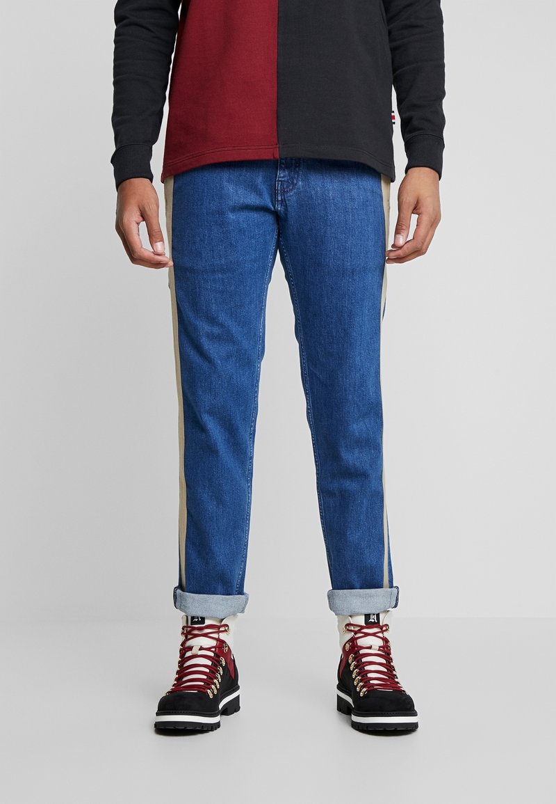 Blå jeans med ljusbeige sidostråk, uppvikta benslut, bärs med svarta vandringsskor med röda snören och guldfärgade detaljer.