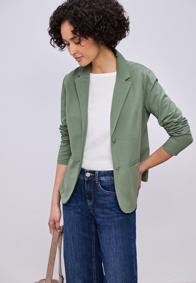 Femme aux cheveux bouclés portant un blazer vert, un haut blanc et un jean bleu, tenant un sac beige d'une main derrière son dos.