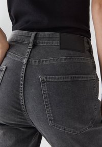 Primo piano di una persona che indossa jeans in denim grigio scuro con tasche posteriori visibili e un'etichetta in pelle nera sulla vita.