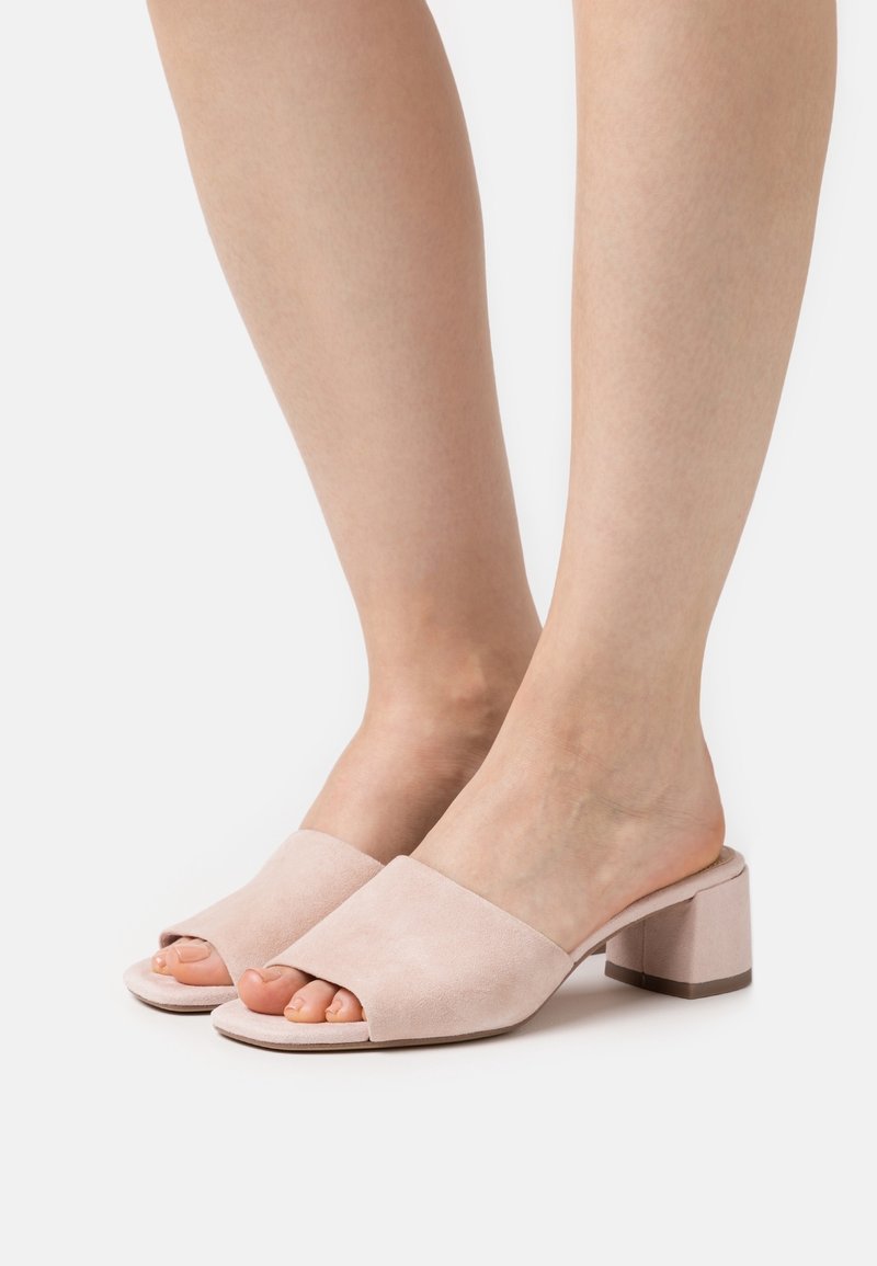 Pieds portant des mules en daim rose clair à bout ouvert avec talon bloc et un design sans bride sur fond blanc uni.