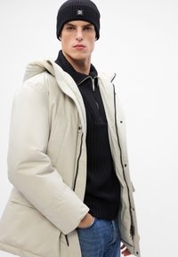 Beige színű pufi dzseki kapucnival, fekete bordázott pulóver alatta, kék farmernadrág, és fekete kötött sapka, elöl cipzárral és gombokkal.