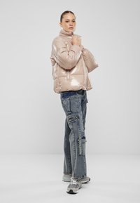 Polstret, glansfull beige jakke med høy krage og glidelåslukking, kombinert med blå cargo-jeans og grå joggesko.