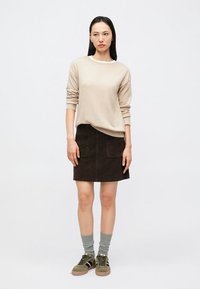 Μπεζ πουλόβερ, σκούρα καφέ σουέντ φούστα με δύο μπροστινές τσέπες, γκρι πλεκτές κάλτσες και πράσινα αθλητικά παπούτσια σε ουδέτερο φόντο.