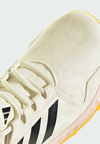 Chaussure de sport beige clair avec un dessus texturé, présentant un motif camouflage subtil, des accents noirs et un design à lacets, plus une semelle jaune.