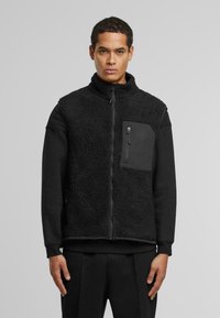 Schwarzener Fleece-Westen mit hohem Kragen, Reißverschluss vorne und Brusttasche. Hat eine strukturierte Oberfläche und schwarze Stoffakzente.
