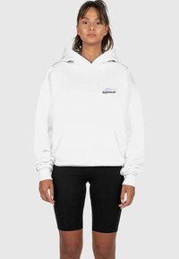 Hvit hettegenser med logo foran, oversized passform, justerbar hette og ribbestrikkede mansjetter, kombinert med svarte sykkelshorts. Glatt stofftekstur.