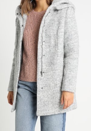 Cappotto di lana grigio con cappuccio, bottoni e cordino. Indossato sopra un maglione di lana rosa, abbinato a jeans azzurri chiari. Texture morbida.