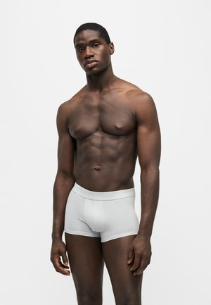 Ein dunkelhäutiges männliches Model ohne Hemd, das hellgraue Boxershorts trägt und vor einem schlichten weißen Hintergrund steht.
