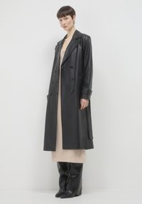 Donna con capelli corti che indossa un lungo cappotto di pelle nera, un vestito beige e stivali neri fino al ginocchio, in piedi su uno sfondo bianco.