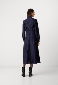 Navyblauwe jurk met lange mouwen, bloemenpatroon, Taille cinch en enkellange zoom. Model draagt zwart hoge hakken.