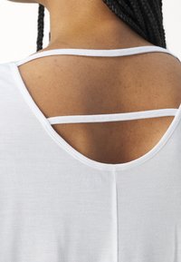 Haut blanc avec une encolure arrondie et un design unique à double bretelle dans le dos, fabriqué à partir d'un tissu doux et lisse.