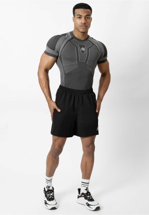Atletische man staat met handen in de zij, draagt een nauwsluitend grijs compressie shirt, zwarte shorts, witte sokken en witte sneakers op een witte achtergrond.