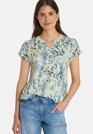 Femme aux longs cheveux foncés portant un chemisier vert clair à manches courtes avec un imprimé abstrait bleu et noir et un jean bleu, une main dans la poche.
