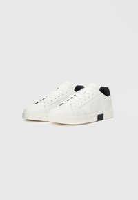 Witte sneakers met geperforeerde zijkanten, platte veters en een zwart accent op de hiel. Gemaakt van synthetisch materiaal met een rubberen zool voor grip.