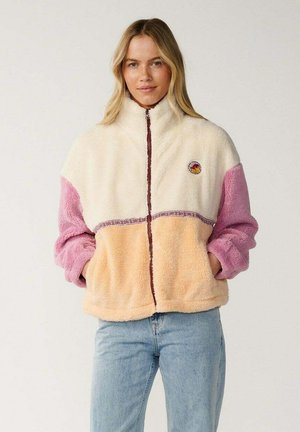 Mujer con una chaqueta de forro polar con bloques de color crema, rosa y melocotón, de pie con las manos en los bolsillos frente a un fondo liso.