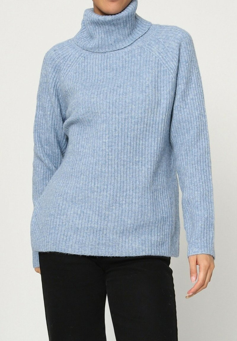 Personne portant un pull col roulé côtelé bleu clair et un pantalon noir, se tenant devant un fond blanc uni.