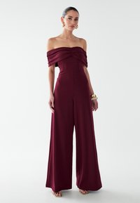 Bordeauxroter Jumpsuit mit Carmenausschnitt, gerafften Details, weitem Bein und glattem Stoff. Akzente setzen goldene Accessoires.