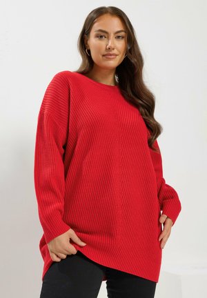 Pull en maille rouge au look décontracté, présentant un motif en côtes texturé, des manches larges et un col rond. Un pantalon noir est visible en dessous.