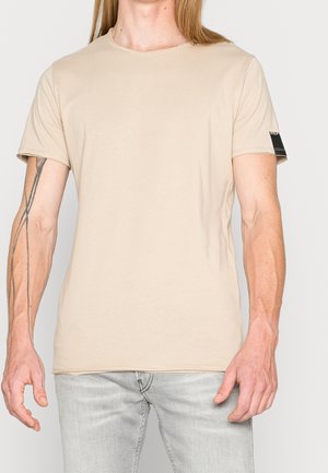 T-shirt beige à manches courtes porté par une personne avec un tatouage sur l'avant-bras gauche, associé à un jean gris clair, debout devant un fond uni.