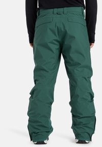 Pantaloni da neve isolati verdi con finitura impermeabile, dotati di una cintura, numerose tasche e fondo con cerniera per una facile vestibilità sopra gli stivali.