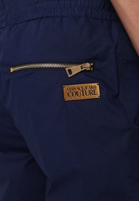 Tumši zila bikses ar redzamu zelta rāvējslēdzēja kabatu un iegravētu zelta uzlīmi ar tekstu "VERSACE JEANS COUTURE." Gluda tekstūra.