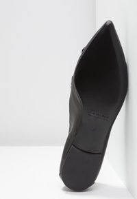 Chaussure en cuir noir à bout pointu avec une texture lisse et une semelle plate. Présente un design minimaliste et un marquage discret sur la semelle.