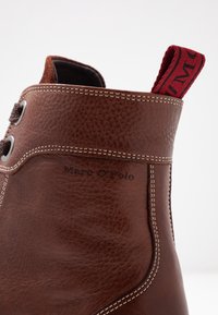 Brun läder ankelsko med en strukturerad yta, vit sömnad, metallöglor och en röd dragrem med svart text.