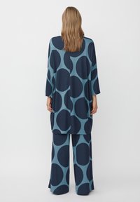 Modrý a tyrkysový vzorovaný dvoudílný outfit, který obsahuje volnou, oversized blůzu s tříčtvrtečními rukávy a široké kalhoty, obojí s velkými kruhy.