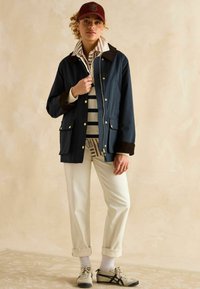 Jachetă marină cu guler din suede maro, accesorii aurii, asortată cu o cămașă în dungi, pantaloni crem și încălțăminte din pânză, pe un fundal neutru.