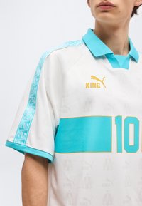 Person iført en hvid og turkis sportstrøje med et gyldent Puma-logo, teksten "KING" og nummer 10 på fronten.