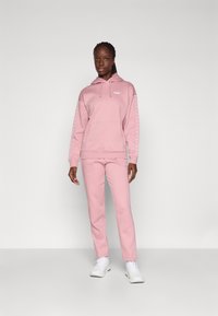 Rosa hettegenser med kengurulomme og snor i hetten, kombinert med matchende rosa joggebukser. Begge har diskrete merkevarelogoer. Hvite joggesko.