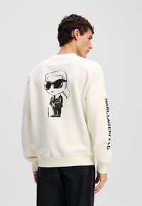 Krémová mikina s potlačou na zadnej strane, ktorá zobrazuje postavu v slnečných okuliaroch, a text "KARL LAGERFELD" pozdĺž rukáva.