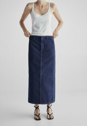 Femme portant un débardeur blanc et une longue jupe en jean bleu foncé avec des sandales noires à brides, debout devant un fond clair uni.