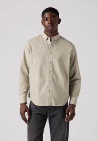 Licht beige overhemd met lange mouwen, een kraag en een subtiel logo. Beschikt over een ontspannen pasvorm en zachte textuur. Gdragen met donkere spijkerbroeken.