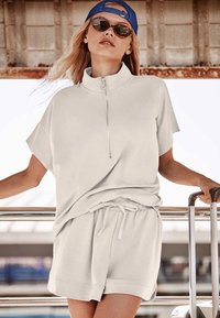 Haut gris clair à fermeture éclair avec manches courtes et short assorti à cordon de serrage. Coupe lisse et décontractée avec un design casual, accessoirisé de lunettes de soleil et d'une casquette bleue.