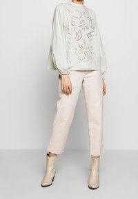 Eine Person trägt eine langärmlige weiße Bluse mit floralen Ausschnitten, helle beigefarbene cropped Hosen und helle beigefarbene Absatzstiefeletten.