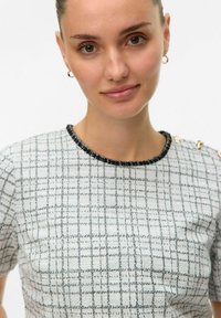Kortärmad topp i ett vävt tyg med ett vit och svart rutig mönster, med en texturerad halsringning och dekorativ hårdvara på axeln.