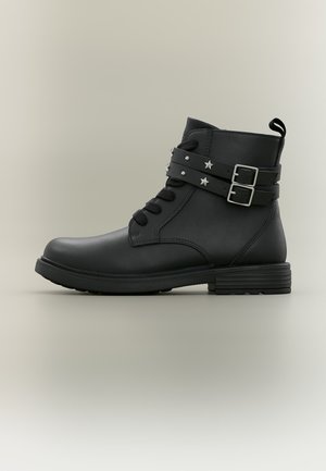 Bottine noire en cuir avec deux sangles décoratives à boucle et clous en forme d’étoile, lacets à l’avant et talon bas à bloc.