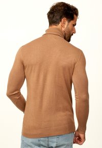 Brauner, gerippter Pullover mit hohem Kragen, tailliertem Schnitt und langen Ärmeln. Der Stoff wirkt weich und leicht und wird mit blauen Jeans kombiniert.