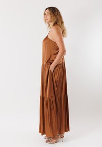 Abito maxi in satin marrone con design a strati e tasche laterali. Caratterizzato da spalline sottili e una silhouette fluida. Abbinato a sandali argentati con cinturini.