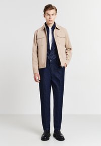 Giacca beige con tasche sul petto, indossata sopra una camicia blu scuro con bottoni e pantaloni di jeans scuri. Stivali neri alla caviglia completano l'outfit.