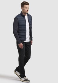 Marineblå puffervest med en glat tekstur, der har lynlås og ribbede marineblå ærmer, båret over en hvid skjorte og sorte jeans.