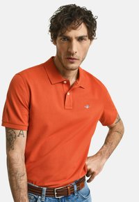 Orange polo t-shirt lavet af bomuld med en klassisk krave, der har et lille logo på brystet, parret med mørke cowboybukser og et brunt bælte.
