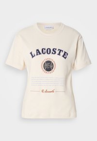 Kremasta pamučna majica s kratkim rukavima, s natpisima "LACOSTE" i "TENNIS HERITAGE" u tamnoplavoj i narančastoj boji, te grafikom krokodila.