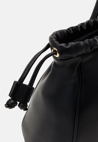 Mustan nahkainen nyörilaukku, jossa on röyhelömäinen yläosa. Varustettu kultasävyisillä metalliosilla ja teksturoidulla, pehmeästi kiiltävällä pinnalla.