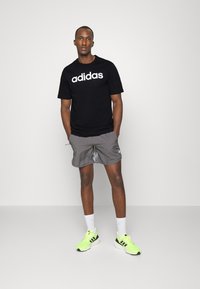 Melna t-krekls ar lielu baltu "adidas" logo, pelēkas sporta šortus, baltas zeķes un neona zaļus sporta apavus ar melniem akcentiem.