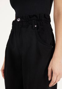 Svarta högmidjade byxor med ett rynkat elastiskt midjeband, framfickor och silverknappdetaljer. Tillverkade av robust bomullstyg.