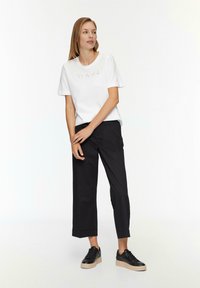 Weißes T-Shirt mit geprägtem "VOYAGE"-Text, kombiniert mit lockeren schwarzen Hosen und schwarzen Sneakers mit einer klobigen beigen Sohle.