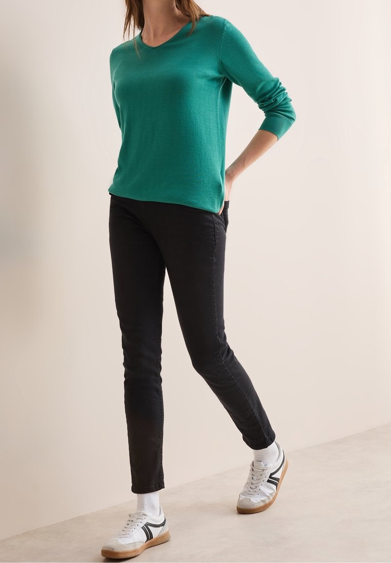 Green knit sweater with a scoop neckline, paired with slim black jeans and white sneakers featuring black stripes. Smooth texture and casual fit.