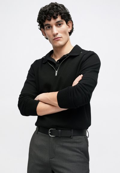Black zip-up sweater with a knitted texture, worn over gray tailored trousers and a black belt featuring a silver buckle, model poses with arms crossed.